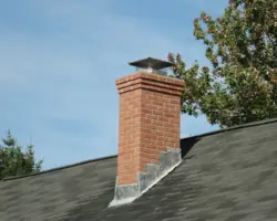 Close-up of a brick chimney stack fitted with a metal cowl and cap against a clear blue sky.