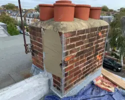 Close-up of a brick chimney stack with four terracotta chimney pots undergoing repair and lead flashing installation on a rooftop.