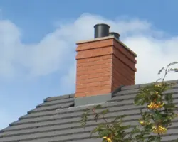 Close-up of a modern brick chimney stack with flashing and leadwork set against a grey tiled roof and blue sky