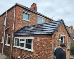 Professional roofing installation on a brick house extension featuring dark tiles, a roof window, and a roofer working below