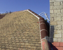 Close-up exterior view of brown interlocking roof tiles, rounded ridge tiles, and custom lead flashing installed around a stone chimney stack under a blue sky.