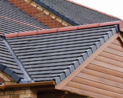 Exterior view of a modern pitched roof featuring dark grey interlocking tiles, contrasting red ridge caps, lead valley flashing, and wood-effect uPVC gable cladding.