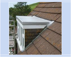Close-up exterior view of a flat-roof dormer window with a smooth lead-topped roof, white frame, and step lead flashing set into brown clay roof tiles.