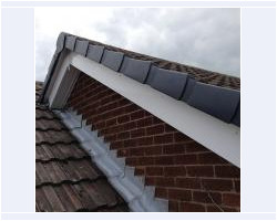 Close-up view of a recently installed grey dry verge roof system, white bargeboards, and step lead flashing on a red brick residential gable wall.