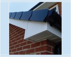 Close-up view of a grey dry verge roof system installed alongside white uPVC fascias, soffits, and black guttering on a red brick property corner.