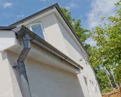 Low-angle view of dark grey PVC guttering and downpipe installed on a modern white rendered residential building with blue sky.
