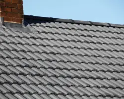 Close-up exterior view of grey interlocking roof tiles with a brick chimney stack and lead flashing set against a clear blue sky.