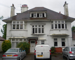 Exterior view of a large detached white house with vehicles parked in front, showing weathered roof tiling and exterior walls needing maintenance