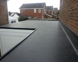 Elevated view of a large, newly installed dark rubber or felt flat roof membrane spanning across a residential extension and garage, running alongside a red brick wall.