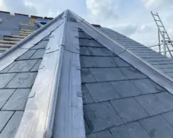 Close-up view of a newly installed lead hip flashing and valleys on a dark slate-tiled roof under a cloudy sky.