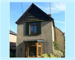 Front view of a pebble-dash two-story house featuring a wooden bay window, dark shingle roof gable, and brown window frames.