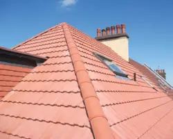 Exterior view of a bright red clay tile hip roof featuring half-round ridge tiles, integrated roof windows, and a tall chimney stack with multiple pots under a clear blue sky