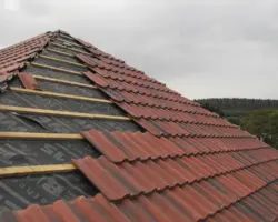 Elevated view of a residential roof undergoing tile installation, showing breathable roofing membrane, wooden battens, and interlocking red clay tiles.