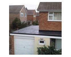 Elevated view of a dark flat roof covering a garage and home extension, connecting to a red brick house with neighboring properties in the background.
