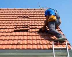Professional roofer working on a bright red clay tile roof using a drill near a ladder and guttering.