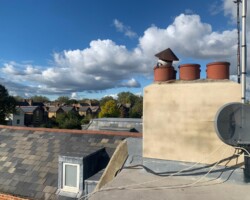 Elevated rooftop view showing a rendered chimney stack with multiple pots, a satellite dish, slate roof tiling, and dormer windows against a blue sky with clouds.