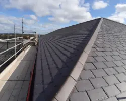 Close-up view of a newly installed dark slate roof with metal capping alongside exterior scaffolding under a cloudy sky.