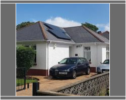 Front view of a white rendered bungalow featuring a dark slate roof with integrated solar panels, a driveway with parked cars, and a low stone boundary wall.