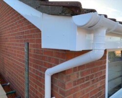 Close-up of freshly installed white uPVC guttering, downpipe, and fascias on the corner of a red brick home with a tiled roof and scaffolding.