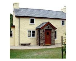 Front view of a yellow-painted country cottage featuring a dark slate roof, dual white chimneys, a stone porch, and a paved patio.