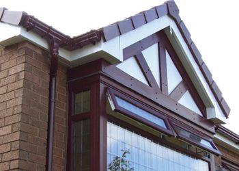 Exterior view of a brown uPVC bay window featuring mock Tudor timber detailing on the gable, matching brown guttering, and a tiled roof edge