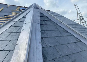 Close-up view of a newly installed lead hip flashing and valleys on a dark slate-tiled roof under a cloudy sky.