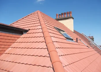 Exterior view of a bright red clay tile hip roof featuring half-round ridge tiles, integrated roof windows, and a tall chimney stack with multiple pots under a clear blue sky