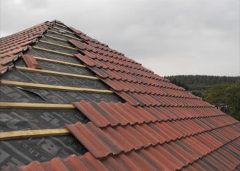 Exterior view of a pitched roof under construction, showing breathable roofing membrane, wooden battens, and interlocking red clay tiles being installed.