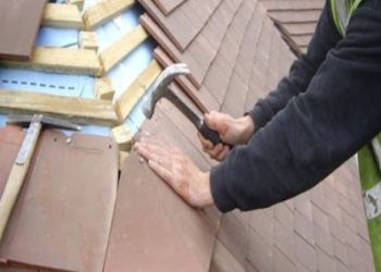 Close-up view of a roofer's hands using a hammer to secure and nail red clay roof tiles onto timber battens over a breathable roofing membrane.