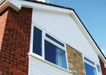 Exterior view of a residential gable wall featuring white uPVC vertical and horizontal cladding, white double-glazed windows, and clean white soffits set against a brick and stone facade under a bright blue sky.